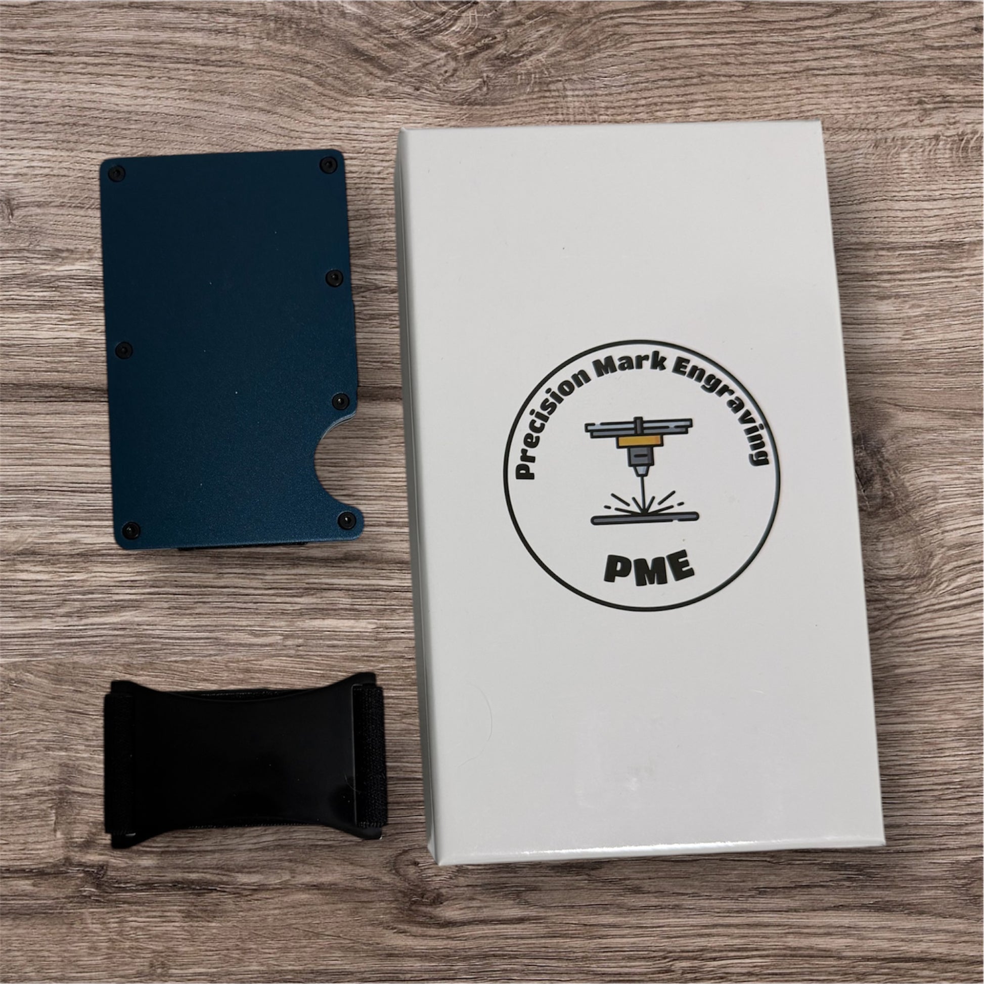 Black metal cardholder and black phone case next to a white box with 'Precision Mark Engraving' logo on a wooden surface.